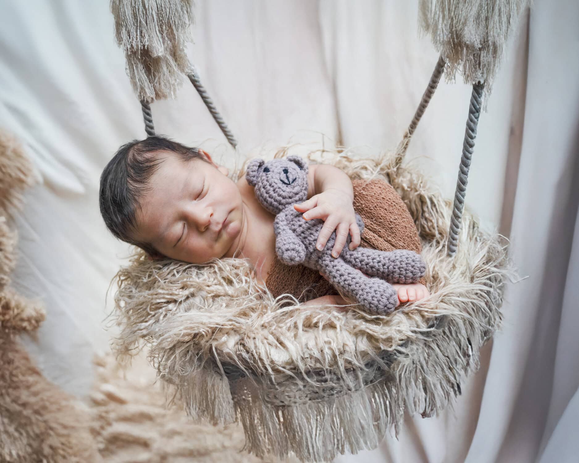 Newborn baby asleep in a hanging fringed basket, holding a small crochet teddy bear, Nook Look studio, Coquitlam