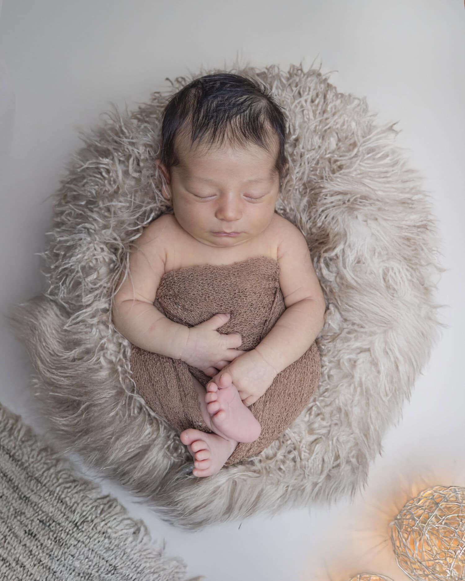 Newborn baby curled up asleep on a cream faux-fur blanket, wrapped in a soft brown swaddle, Nook Look studio