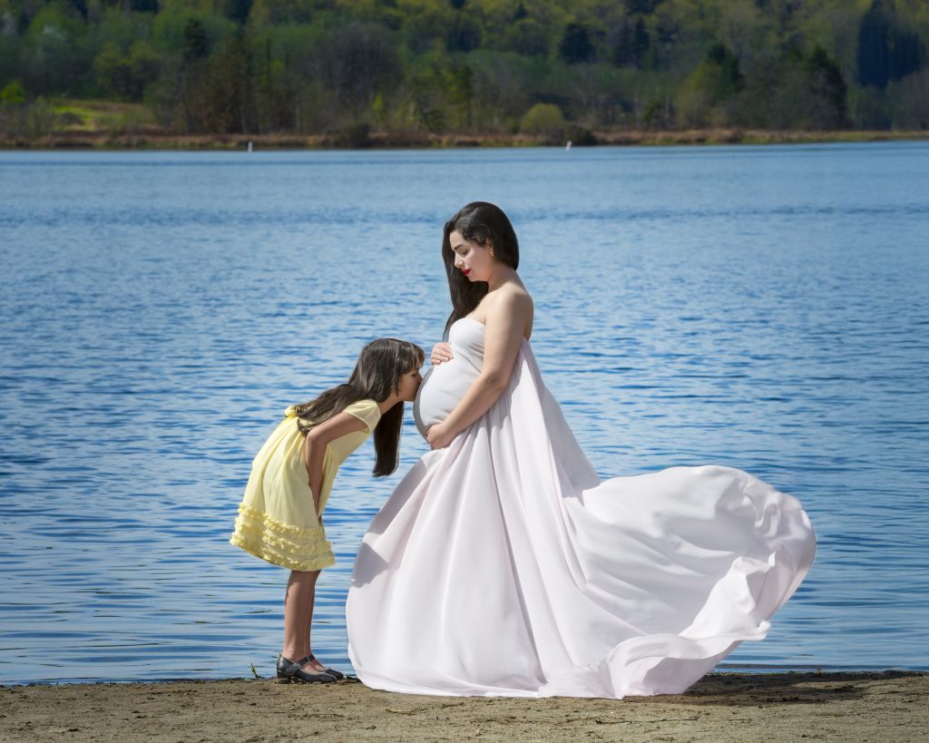 Pregnant mother in a flowing white gown by the lake, her young daughter kissing her belly, Nook Look outdoor maternity session