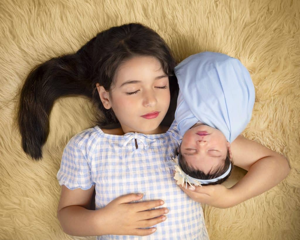 Older sister cuddling her newborn sibling, who wears a blue knit wrap and floral headband, on a fur blanket, Nook Look studio